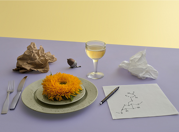 Table set, there is a flower on a plate. Alongside fork, knife, glass there is a noteblock and a pen. On the notebook there is a chemical formula.